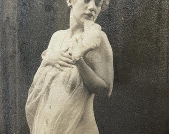 Può includere: Fotografia vintage in bianco e nero di una persona, parzialmente drappeggiata in un tessuto trasparente. La persona ha la pelle chiara con i capelli ricci ed è in posa in stile classico. Lo sfondo è una superficie scura e strutturata.