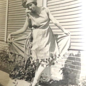 May include: A woman in a white dress with a decorative trim stands in front of a wooden slat wall. She is wearing a hat with a flower design.