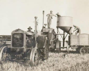 Può includere: Fotografia in bianco e nero di un trattore e attrezzature agricole d'epoca in un campo. Diverse persone caricano grano su un rimorchio. L'immagine cattura un momento della storia agricola.
