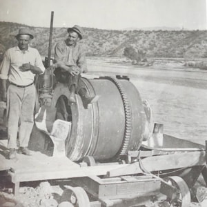 May include: Two men stand next to a vintage concrete mixer on a wooden platform. The mixer has a large drum and a smaller hopper. The men are wearing hats and work clothes. The mixer is on a dirt road.