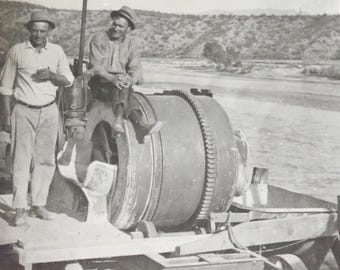Può includere: Due uomini stanno in piedi accanto a una betoniera vintage su una piattaforma di legno. La betoniera ha un grande tamburo e una tramoggia più piccola. Gli uomini indossano cappelli e abiti da lavoro. La betoniera è su una strada sterrata.