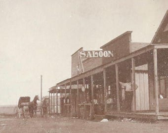 Può includere: Una fotografia in bianco e nero di un saloon in una città del West. Il saloon ha un portico in legno con un cartello che recita "SALOON". Ci sono persone in piedi sul portico e una carrozza trainata da cavalli davanti al saloon.