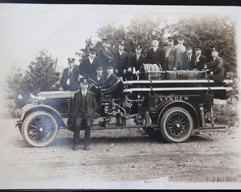 Può includere: Una fotografia d'epoca in bianco e nero di un gruppo di uomini in piedi davanti a un camion dei pompieri. Il camion è etichettato "Lynden" e ha un grande tubo sul retro. Gli uomini indossano abiti e cappelli. La foto sembra essere degli inizi del 1900.