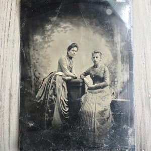May include: A black and white vintage photograph of two women in dresses posing for a portrait. The women are standing next to each other and are looking directly at the camera. The woman on the left is wearing a long, dark dress with a white collar and a bow. The woman on the right is wearing a lighter dress with a lace collar and a bow. Both women have their hair pulled back and are wearing simple jewelry.