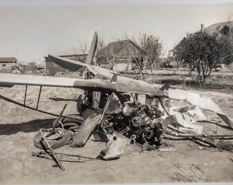 A vintage airplane lies wrecked and crumpled on a dirt ground, surrounded by debris, with a backdrop of trees and houses, suggesting a crash site.
