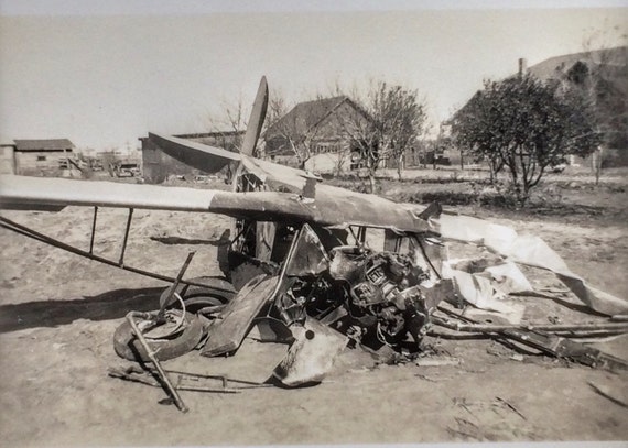A vintage airplane with a severely damaged fuselage and broken wings lies wrecked on the ground, surrounded by debris, with houses and trees in the background.