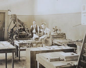 Può includere: Una foto vintage in bianco e nero di una macchina da stampa in un laboratorio. Due persone stanno lavorando alla macchina da stampa, e ci sono pile di carta su tavoli sullo sfondo.
