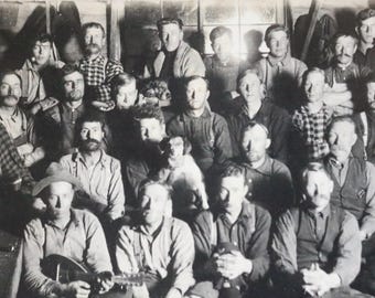 Può includere: Una fotografia in bianco e nero di un gruppo di uomini in piedi insieme. Alcuni degli uomini indossano cappelli e alcuni tengono degli strumenti. C'è un cane nella foto.
