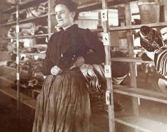 May include: A woman stands in a shop, looking to the right. She is wearing a dark blouse and a long skirt. She is standing in front of a shelf with various items on it. The shelf is labeled with numbers.