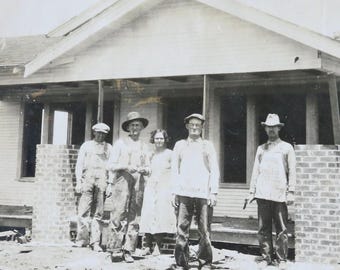 Può includere: Una fotografia in bianco e nero di un gruppo di persone in piedi davanti a una casa in costruzione. Ci sono cinque persone nella foto, tre uomini e due donne. Gli uomini indossano cappelli e le donne indossano abiti. La casa è in legno e ha un portico.