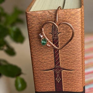 Peut inclure: Un marque-page en fil de cuivre en forme de cœur, doté d'un crochet qui se glisse sur la page d'un livre. Le fil est orné de petites perles translucides roses et vertes. Le marque-page est placé sur la tranche d'un livre brun avec un motif d'épée.