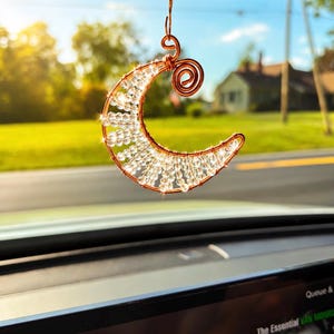 May include: A crescent moon ornament crafted from copper-colored wire and clear faceted beads. The moon shape features a spiral wire detail at the top and is suspended by a matching copper-colored wire hook, catching the sunlight.