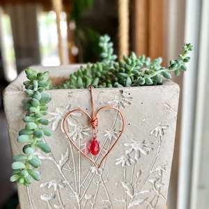 May include: A copper wire heart ornament featuring a single faceted red teardrop bead suspended from the center. The heart is shaped from a continuous piece of wire with a wrapped coil at the top, creating a simple and delicate decorative accent piece.