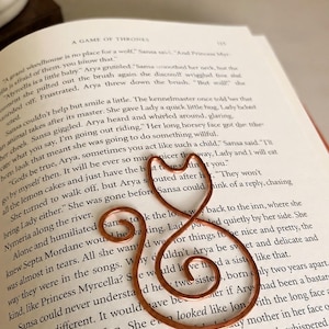 May include: A copper-colored wire bookmark shaped like a cat silhouette with a curled tail and pointed ears, resting on an open page of the book A Game of Thrones. The metal wire is bent into a continuous outline forming the head and body of a cat.