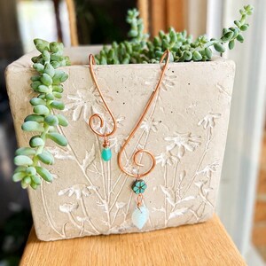 May include: A copper wire ornament featuring two spiral loops, each adorned with a small dangling bead. One loop holds a single bright turquoise teardrop bead, while the other features a small flower-shaped bead above a pale blue teardrop bead.