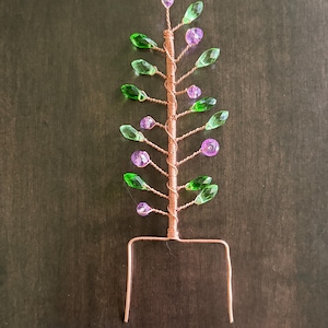 May include: A decorative wire sculpture shaped like a small tree, crafted from twisted copper wire. The branches are adorned with faceted, teardrop-shaped green beads and round, translucent purple beads, ending in a U-shaped base for insertion.