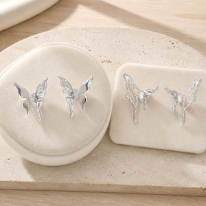 May include: Two pairs of silver-toned butterfly earrings. The first pair features solid, smooth wings with a central cluster of small clear stones. The second pair has an open, fluid, wire-like wing design with a single clear stone at the centre.