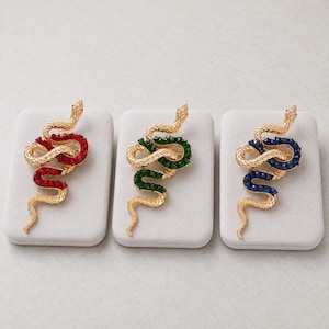 May include: Three gold-toned snake brooches, each featuring a textured metallic body coiled into a serpentine shape. One brooch is accented with red crystals, one with green crystals, and one with blue crystals along the snake's back.
