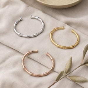 May include: Three open cuff bracelets designed to resemble bamboo stalks. One bracelet is silver-toned, one is gold-toned, and one is rose gold-toned. Each piece features a segmented, cylindrical structure with a smooth, polished metallic finish.