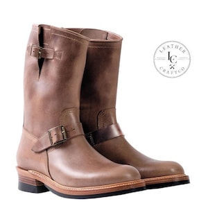 May include: A pair of mid-calf brown leather boots featuring a rounded toe, a stacked heel, and a dark brown rubber outsole. Each boot includes an adjustable strap with a metal buckle across the instep and a smaller buckle at the top collar.