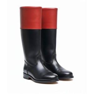 May include: A pair of knee-high boots featuring a smooth black leather lower section and a contrasting solid red leather cuff at the top. The boots have a rounded toe, a low block heel, and a structured shaft that reaches just below the knee.