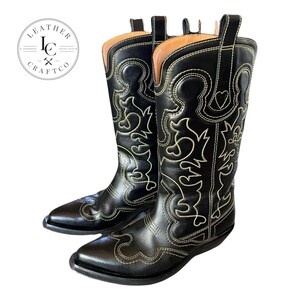 May include: A pair of black leather cowboy boots featuring intricate light-coloured decorative stitching patterns across the shaft and foot. The boots have a pointed toe, a stacked heel, and pull tabs at the top. A circular logo reads LEATHER CRAFTCO.