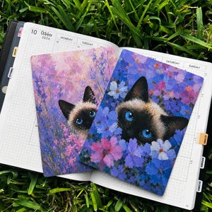 May include: Two rectangular prints featuring Siamese cats with bright blue eyes peeking through vibrant purple, pink, and blue floral backgrounds, resting on an open planner with the text 10 October 2026 visible at the top of the page.