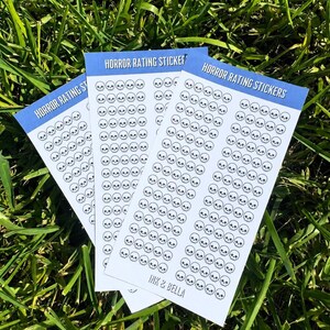 May include: Three rectangular sheets of white stickers featuring rows of small black skull icons. Each sheet has a blue header with the text HORROR RATING STICKERS and the brand name INK & BELLA printed in black at the bottom of the white section.