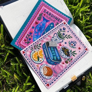 May include: Two rectangular postcards featuring colorful illustrations of sardines. One postcard has a pink background with blue fish and the text SARDINES in olive oil. The other shows a tin of sardines, orange slices, bread, and olives.