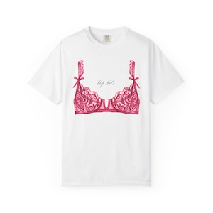 Può includere: Una t-shirt bianca a maniche corte con la grafica di un reggiseno in pizzo rosa con piccoli fiocchi sulle spalline. Il testo "big hits" è stampato in un carattere corsivo nero centrato sopra l'illustrazione del reggiseno sul davanti.