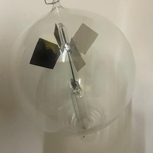 May include: A clear glass radiometer featuring a spherical bulb with a central vertical glass stem. Inside, a delicate rotor holds four thin, square vanes, each with one dark side and one light side, suspended on a needle point for rotation.