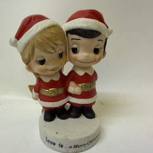 May include: A ceramic figurine of two children wearing red and white Santa hats and matching red outfits with gold belts. They stand on a circular white base with the text "love is...a Merry Christmas" printed in black on the front of the base.