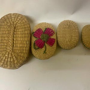 May include: A set of four oval-shaped woven baskets made from natural tan fibers. The baskets vary in size, with the second smallest featuring a decorative pink and green flower design stitched onto the lid. Each basket has a matching woven lid.