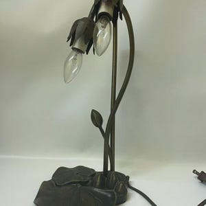 May include: A bronze-toned metal table lamp featuring two light bulbs encased in petal-shaped shades. The lamp base is designed with flat, circular lily pad shapes, and the stems curve upward to hold the light fixtures, complete with a power cord.