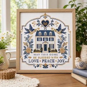 Puede incluir: Un bordado en punto de cruz enmarcado que presenta una ilustraci&oacute;n de una casa, dos p&aacute;jaros, motivos florales y el texto "MAY THIS HOME BE BLESSED WITH LOVE PEACE JOY" en hilo azul y dorado sobre tela blanca en un marco de madera clara.