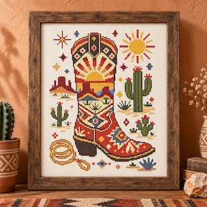 May include: A framed cross-stitch embroidery featuring a central red and yellow cowboy boot decorated with geometric patterns. The design includes a yellow sun, green cacti, a coiled rope, and small star motifs on a white fabric background.