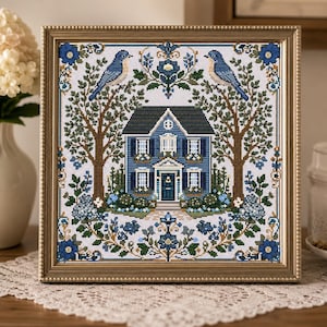 May include: A framed cross-stitch embroidery featuring a blue house centred between two trees with blue birds perched on the branches. The design includes floral motifs, green foliage, and decorative borders in shades of blue, brown, and green on a white fabric background.