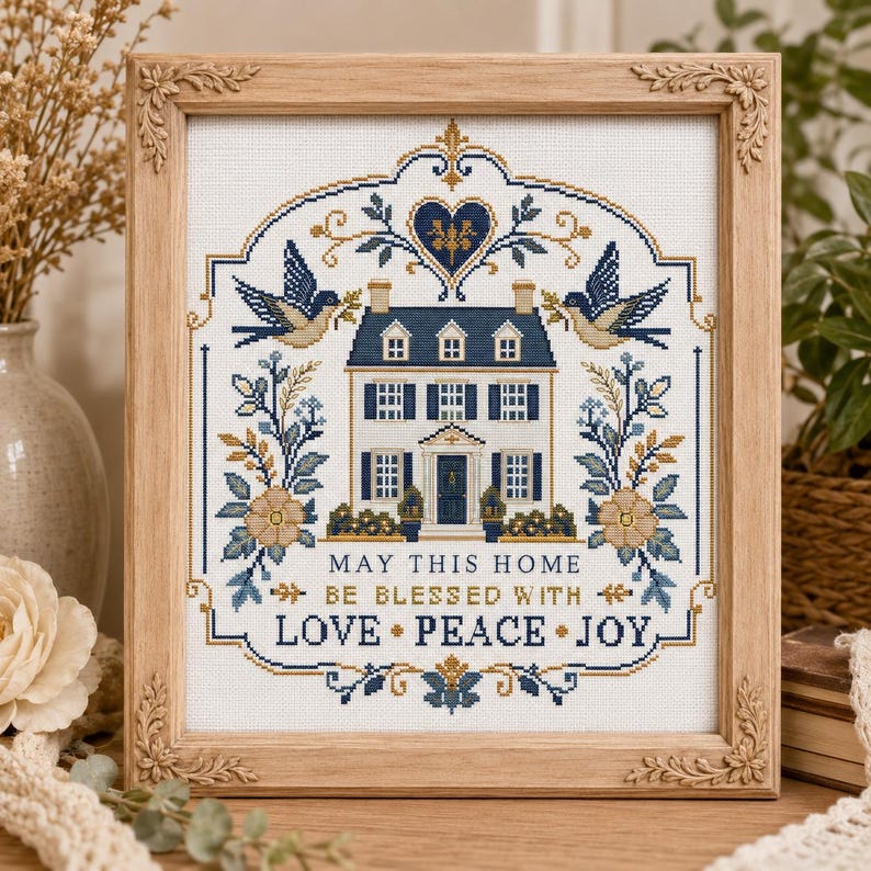 Puede incluir: Un bordado en punto de cruz enmarcado que presenta una casa central con contraventanas azules, flanqueada por p&aacute;jaros y motivos florales. El texto dice: MAY THIS HOME BE BLESSED WITH LOVE PEACE JOY. Dise&ntilde;o con hilos azul, dorado y blanco sobre tela blanca.