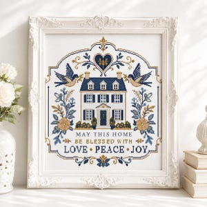 Puede incluir: Un bordado en punto de cruz enmarcado que presenta una ilustraci&oacute;n central de una casa rodeada de p&aacute;jaros, motivos florales y un coraz&oacute;n. El texto dice: MAY THIS HOME BE BLESSED WITH LOVE PEACE JOY. El dise&ntilde;o usa una paleta de azul marino, oro y blanco.