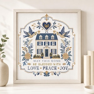 May include: A framed cross-stitch embroidery featuring a central house illustration, two birds, floral motifs, and the text: MAY THIS HOME BE BLESSED WITH LOVE • PEACE • JOY. The design uses a color palette of navy blue, tan, and white on fabric.