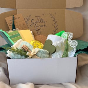 May include: A gift box containing a reading list card, green pens, a yellow notebook, a round cork coaster, a small yellow candle tin, a green heart-shaped item, a white mug, a green fuzzy cloth, a wooden honey dipper, and a clear bottle-shaped tag.