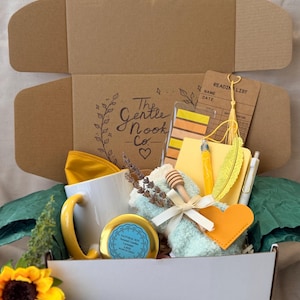 May include: A gift box containing a white mug with a yellow handle, a round candle tin, a light blue fuzzy item with a wooden honey dipper, a yellow notebook, a yellow feather bookmark, a reading list card, and a pen, with The Gentle Nook Co. text.
