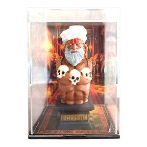 Può includere: Un busto del personaggio Dhalsim esposto all'interno di una teca in acrilico trasparente. La figura presenta un turbante bianco, una lunga barba bianca, segni rossi sul viso e una collana di tre teschi bianchi su una base nera con il nome Dhalsim.