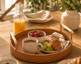 May include: A round woven rattan serving tray with two light-colored wooden handles, containing a ceramic mug filled with coffee, a small bowl of fresh strawberries, a plate with a croissant and green grapes, and a glass of orange juice.