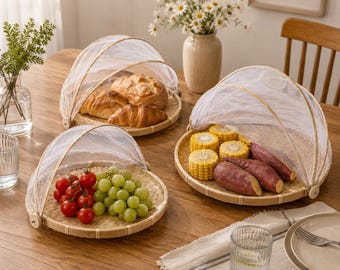 May include: Three round woven bamboo trays with white mesh dome covers. One tray holds croissants, another holds green grapes and cherry tomatoes, and the third holds corn on the cob and sweet potatoes. The trays sit on a wooden table.