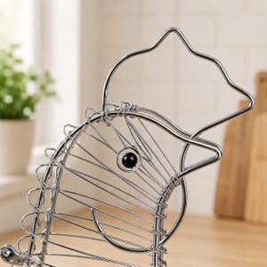 May include: A silver-colored wire basket shaped like a chicken, constructed from thin metal rods. The structure features a prominent wire outline of a comb and beak, with a single black bead serving as the eye, and a coiled wire body design.
