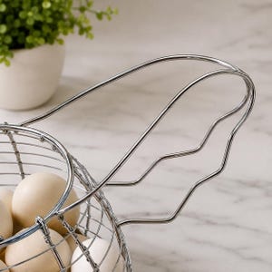 May include: A silver-colored wire egg basket featuring a rounded base and a prominent, wavy-edged handle. The basket is filled with several smooth, off-white eggs, showcasing a metallic construction with a polished finish and a grid-like design.