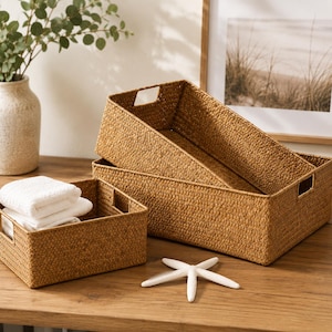 May include: Two rectangular woven storage baskets made of natural tan fibers, featuring integrated side handles. A stack of folded white towels sits inside the smaller basket, and a white starfish is placed on the surface in front of them.