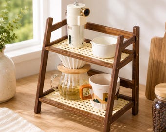 May include: A two-tiered wooden shelf with a light-colored woven cane surface on each level. The top shelf holds a white coffee grinder and a small white pitcher. The bottom shelf holds a glass coffee carafe and a white mug with a brown handle.