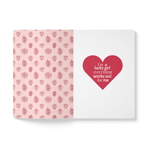 Puede incluir: Un cuaderno rectangular con un diseño de cubierta dividido. El lado izquierdo presenta un fondo rosa claro con un patrón repetitivo de pequeños iconos de corazones rojos. El lado derecho es blanco con un gran corazón rojo que contiene el texto I'm a lucky girl everything works out for me.